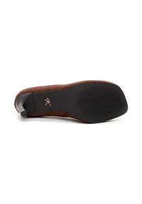Zapato de tacón alto de ante marrón con una silueta curvada, que presenta una suela de goma negra con un patrón de relieve geométrico y un acento dorado.