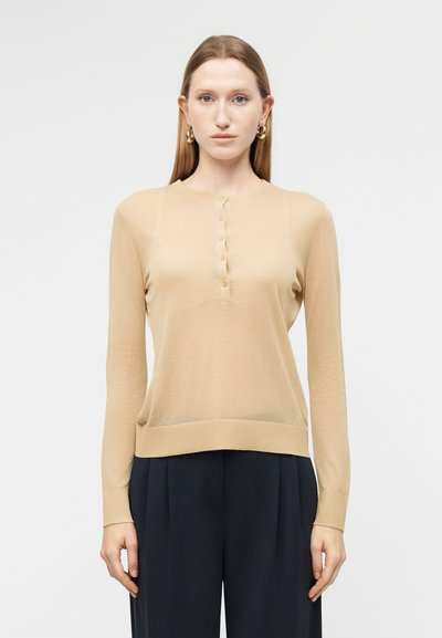 Femme portant un haut beige à manches longues boutonné et un pantalon noir, debout devant un fond clair uni, face à l'avant.