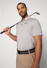 Pelēks polo krekls ar pogu plāksteri un melnu Nike logotipu, apvienots ar bežām biksēm un melni svītrotu jostu; tur golfu nūju.