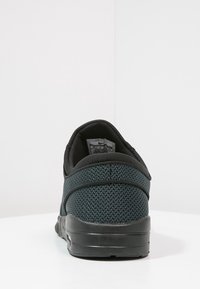 Svart sneaker med en strukturerad mesh-överkonstruktion, vadderad krage och en slät svart gummisula. Utmärkande sömnadsdetaljer på hälområdet.