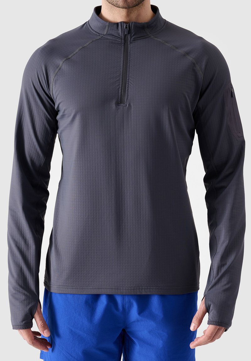 Dunkelgraues Langarm-Pullover-Shirt mit einem Reißverschluss in halber Länge, strukturiertem Stoff und einer Reißverschlusstasche am linken Arm. Darunter werden blaue Shorts getragen.