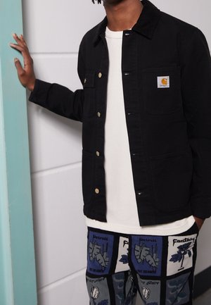 Personne portant une veste noire sur une chemise blanche et un pantalon à motifs, une main reposant sur un encadrement de porte bleu clair.