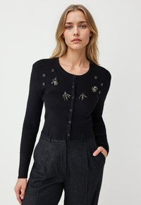 Cardigan noir à côtes avec manches longues, arborant une broderie décorative et six boutons à l'avant. Porté avec un pantalon tailleur sombre.