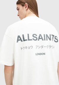 White cotton t-shirt with oversized fit, featuring bold black text "ALLSAINTS" and additional text in Japanese and "LONDON" on the back.