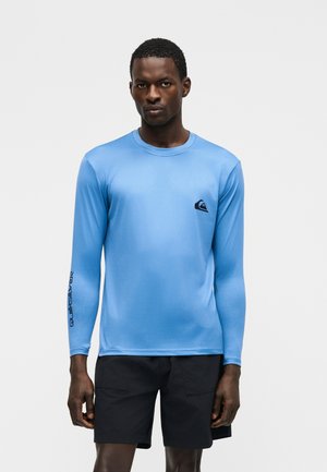 Άνδρας που φοράει μπλε ριπλό μπλουζάκι με μακριά μανίκια και μαύρο λογότυπο στο στήθος και στον μανίκι, σε συνδυασμό με μαύρο μαγιό, στέκεται μπροστά σε λευκό φόντο.