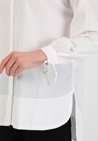 Witte blouse met knoopsluiting aan de voorkant, lange pofmouwen en gathered manchetten vastgebonden met een lint. Gemaakt van een lichtgewicht, iets doorschijnende stof.