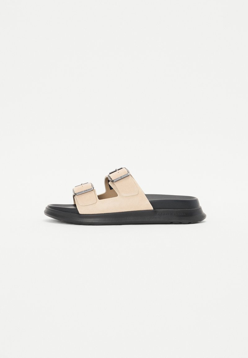 Beige semskede sandaler med to justerbare stropper og sølvspenner, hvilende på en sort gummisåle. Glatt overflate med minimal tekstur.