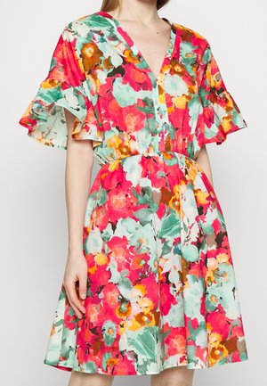 Žena nosí krátké šaty ke kolenům s živým květinovým vzorem v červené, oranžové, zelené a bílé, s volánovými rukávy a výstřihem do V.