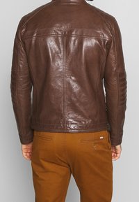 Hnědá kožená bunda s hladkou texturou, prošívanými ramenními akcenty a přiléhavým střihem. V kombinaci s béžovými kalhotami s zadními kapsami.