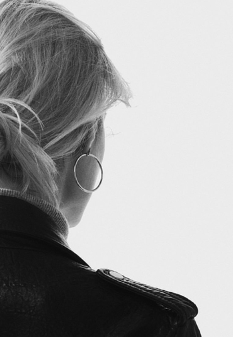 Femme aux cheveux blonds en chignon bas, portant de grandes boucles d’oreilles créoles et une veste en cuir foncée, dos tourné sur un fond clair uni.