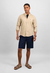 Camicia beige con colletto a band, pantaloni corti blu navy e sandali marroni. Tessuto morbido, design semplice e un piccolo logo sulla tasca della camicia.