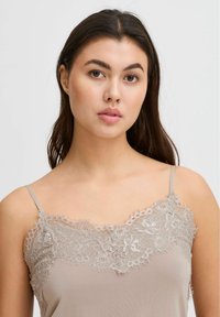 Camiseta sin mangas beige con un delicado encaje en el escote, con tirantes finos y una tela suave y lisa. El encaje incluye patrones florales.