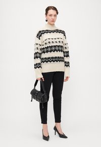 Strickpullover mit geometrischen Mustern in Schwarz und Creme, geripptem Kragen und Saum, kombiniert mit schwarzen Skinny-Hosen und einer kleinen schwarzen Tasche.
