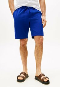 Pantaloni da bagno blu realizzati in materiale leggero, con vita elastica e cordoncino e tasche laterali. Indossati con sandali scivolati marroni.