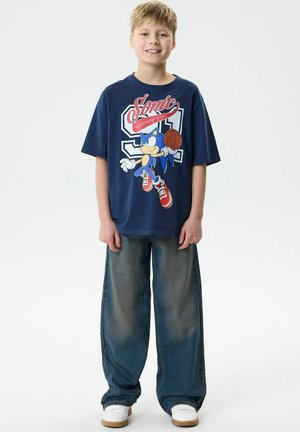 Garçon souriant portant un t-shirt graphique de basketball Sonic the Hedgehog bleu marine, un jean bleu à jambes larges et des baskets blanches, debout devant un fond uni.