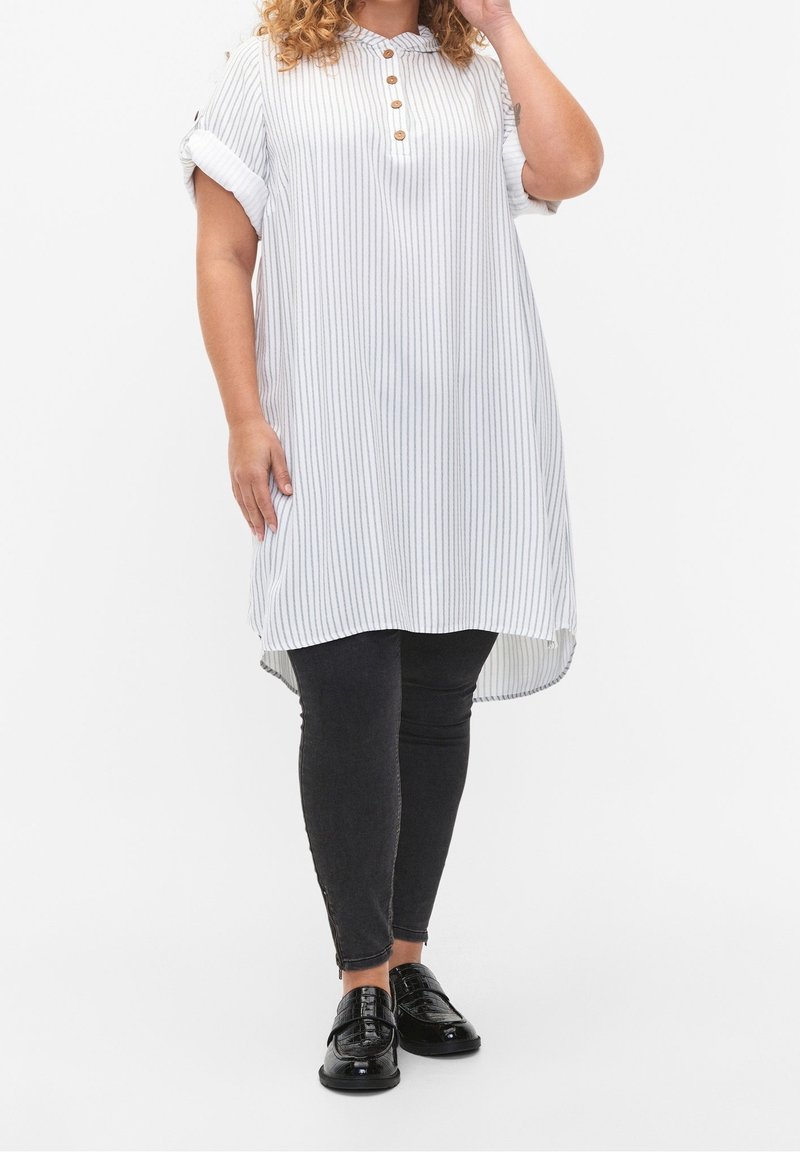 Tunique rayée blanche avec col boutonné, manches courtes retroussées et ourlet asymétrique, associée à un jean skinny noir et des chaussures noires.