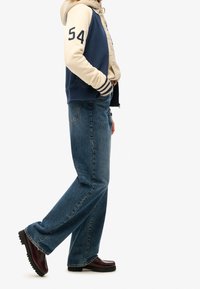 Granatowo-kremowa bluza z kapturem z numerem „54”, zestawiona z niebieskimi prostymi jeansami i ciemnymi skórzanymi butami o teksturowanej podeszwie.