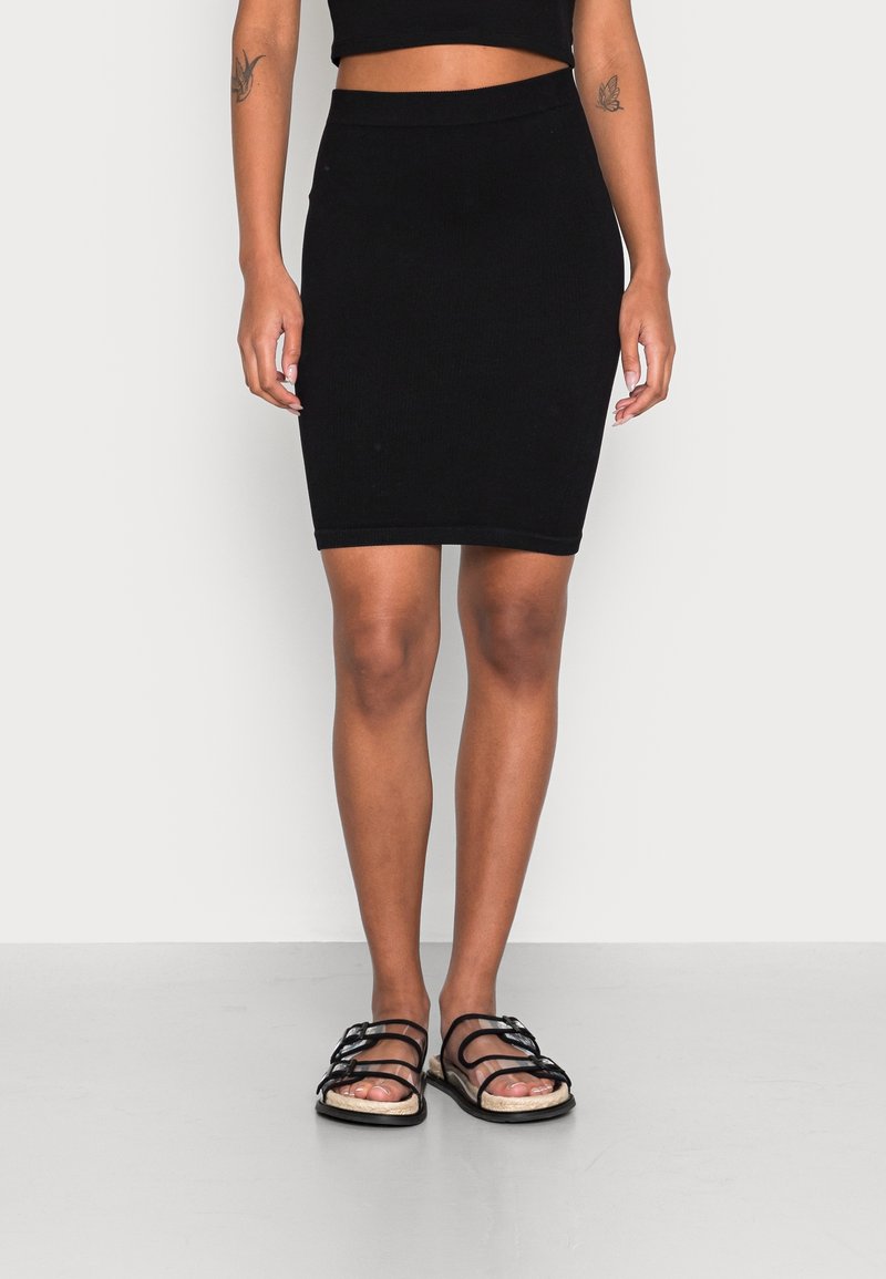 Svart ribbad midijupe med en åtsittande design, med ett brett midjeband, tillsammans med svarta sandaler med remmar på ett ljusgrått golv.