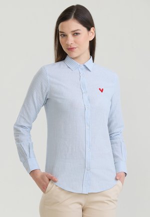 Jeune femme debout les mains dans les poches, portant une chemise à rayures bleu clair avec des boutons et un petit cœur rouge sur la poitrine.