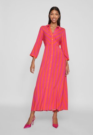 Uśmiechnięta kobieta, stojąca, ubrana w długą sukienkę zapinaną na guziki z pionowymi różowo-pomarańczowymi paskami i różowymi szpilkami.