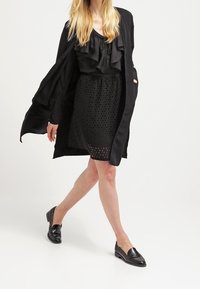 Blusa nera con ruches abbinata a una gonna nera con taglio laser. L'outfit è completato da mocassini neri e un lungo cappotto nero aperto.