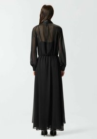 Lungo abito nero realizzato in tessuto trasparente con maniche lunghe, vita aderente e gonna fluente, caratterizzato da un colletto alto e una scollatura sulla schiena.