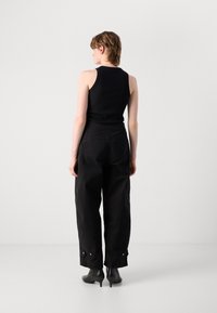 Persona in piedi con la schiena rivolta, che indossa un top senza maniche nero, pantaloni larghi neri e stivaletti con tacco neri su uno sfondo neutro.