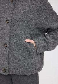 Gray wool coat with large buttons and a patch pocket. Features a loose fit and textured fabric, suitable for cool weather.