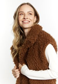 Brun, fluffy vest med en tekstureret, pelslignende overflade. Har en åben front, en bred krave og bæres over en hvid langærmet bluse.
