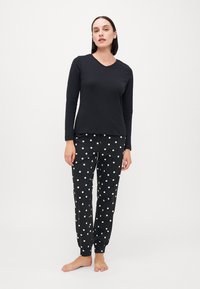 Top negru cu mânecă lungă și decolteu în v, asociat cu pantaloni de pijama negri cu buline albe și manșete elastice la glezne.