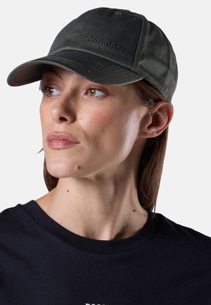 Donna che indossa un berretto da baseball Rossignol verde scuro e una maglietta nera, che guarda verso sinistra contro uno sfondo chiaro e semplice.