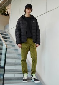 Black puffer jacket, black long-sleeve shirt, olive green cargo pants, and black sneakers with neon accents. Standing on stairs inside a building.