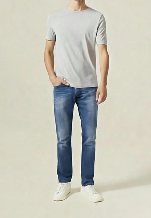 Mann trägt ein hellgraues T-Shirt mit kurzen Ärmeln, blaue Jeans und weiße Turnschuhe, steht mit einer Hand in der Tasche vor schlichtem Hintergrund.