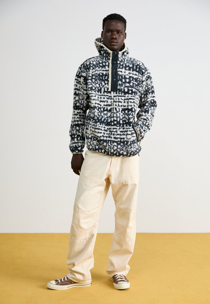 Polarowy sweter z wysokim kołnierzem, w czarno-białym wzorze, z krótkim zamkiem i kieszeniami bocznymi, w zestawieniu z jasnobeżowymi spodniami i brązowymi trampkami.