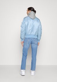 Man met donker haar in een knot, draagt een lichtblauwe bomberjack, grijze hoodie, blauwe spijkerbroek en witte sneakers, staand met zijn rug naar de kijker.