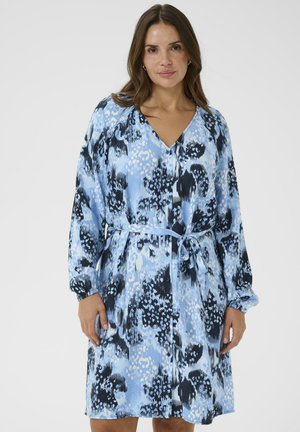 Femme portant une robe à manches longues à motifs abstraits bleus et noirs, avec un col en V et une ceinture nouée à la taille, debout devant un fond uni.