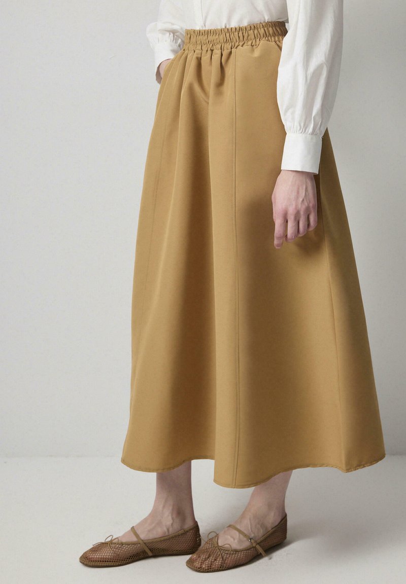 Una falda mostaza de longitud media con una cintura elástica, que presenta costuras verticales y una tela suave y texturizada. Combinada con unos bailes color beige.