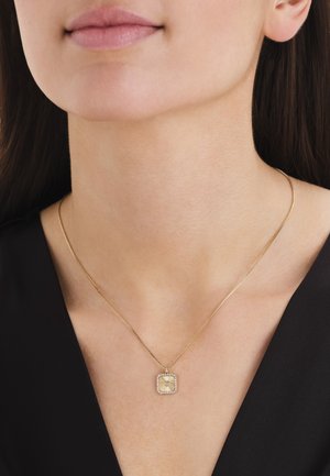 Close-up of a person wearing a thin gold chain necklace with a square gold pendant adorned with small clear stones.
