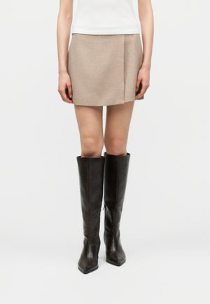 Person iført en beige mininederdel og sorte knæhøje læderstøvler mod en ensartet hvid baggrund.