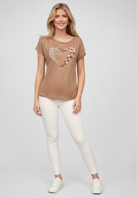 Brun t-skjorte med kort erme og design av et hjerte og sommerfugl i forskjellige farger, kombinert med hvite trange jeans og hvite joggesko.