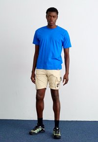 Tricou din bumbac albastru, pantaloni scurți cargo bej cu buzunare, șosete negre și pantofi sport negri-verzi. Cumpănind pe un fundal alb simplu.