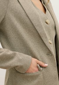 Blazer a spina di pesce di colore marrone chiaro con texture a contrasto, chiusura con un bottone e tasca sulla manica sinistra. Tessuto morbido e vestibilità su misura.