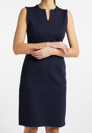 Granatowa sukienka bez rękawów, do kolan, z dekoltem w kształcie litery V, noszona przez osobę stojącą z rękami skrzyżowanymi na wysokości pasa.