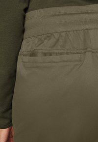 Pantalon vert olive avec une taille élastique, comportant une poche arrière. Le tissu est lisse avec un léger éclat, et le design est ajusté.