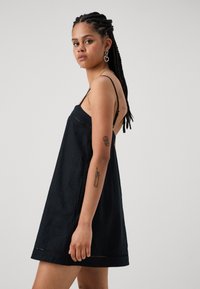 Schwarzes Leinenkleid mit lockerer Passform, dünnen Trägern und seitlichen Schlitzen. Verfügt über feine Nähte entlang dem Saum und eine glatte Textur.