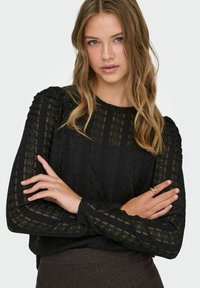 Svart bluse i lacet med lange ermer og intrikate mønstre, rund utringning og teksturert stoff. Modellen har armene i kors.