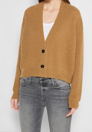 Femme portant un cardigan en maille camel boutonné par-dessus un haut blanc, associé à un jean gris délavé usé.