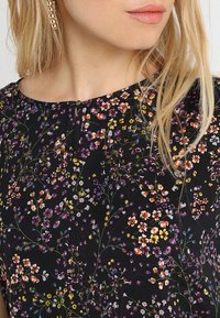 Blusa negra con estampado floral multicolor que incluye flores en tonos morado, rosa y amarillo. Presenta un cuello redondo y detalles de botones.