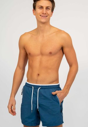 Glimlachende shirtloze man met een blauwe zwembroek met witte aansnoeringskoorden, staand met één hand in de zak tegen een effen achtergrond.