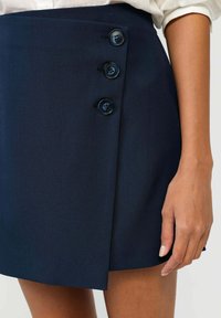 Jupe portefeuille bleu marine avec quatre boutons sur le côté gauche, fabriquée en tissu lisse, présentant un design épuré et structuré avec une fente sur le côté.